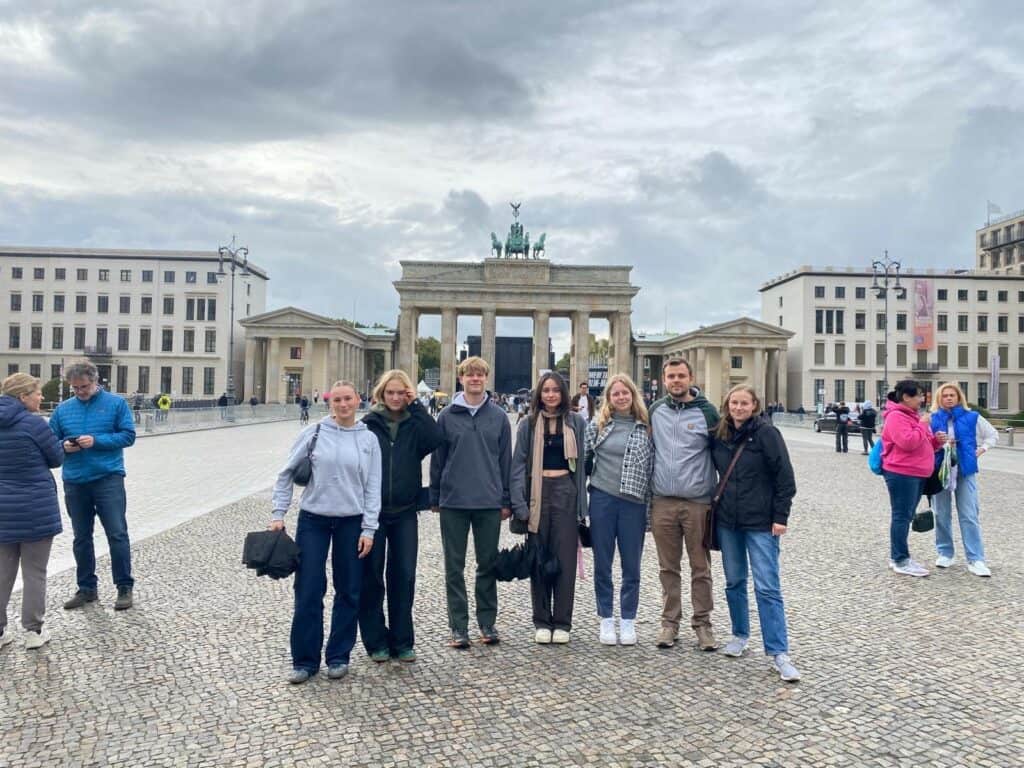 Eine Gruppe von sieben Personen steht unter wolkenverhangenem Himmel vor dem Brandenburger Tor.