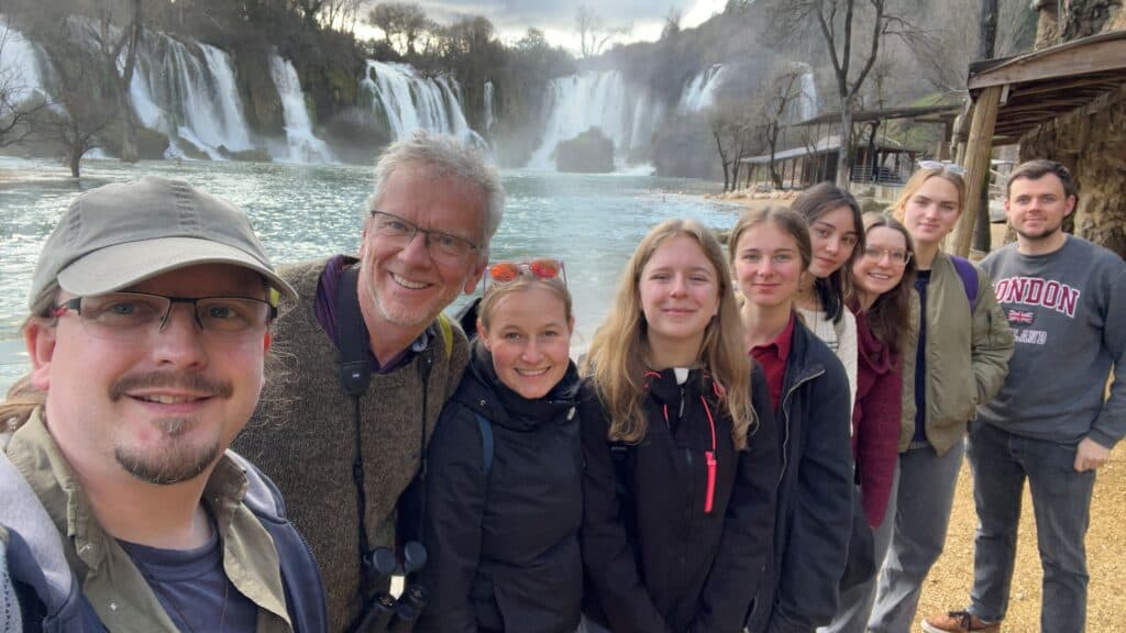 Eine Gruppe von Menschen steht tagsüber lächelnd vor einer malerischen Wasserfallkulisse.