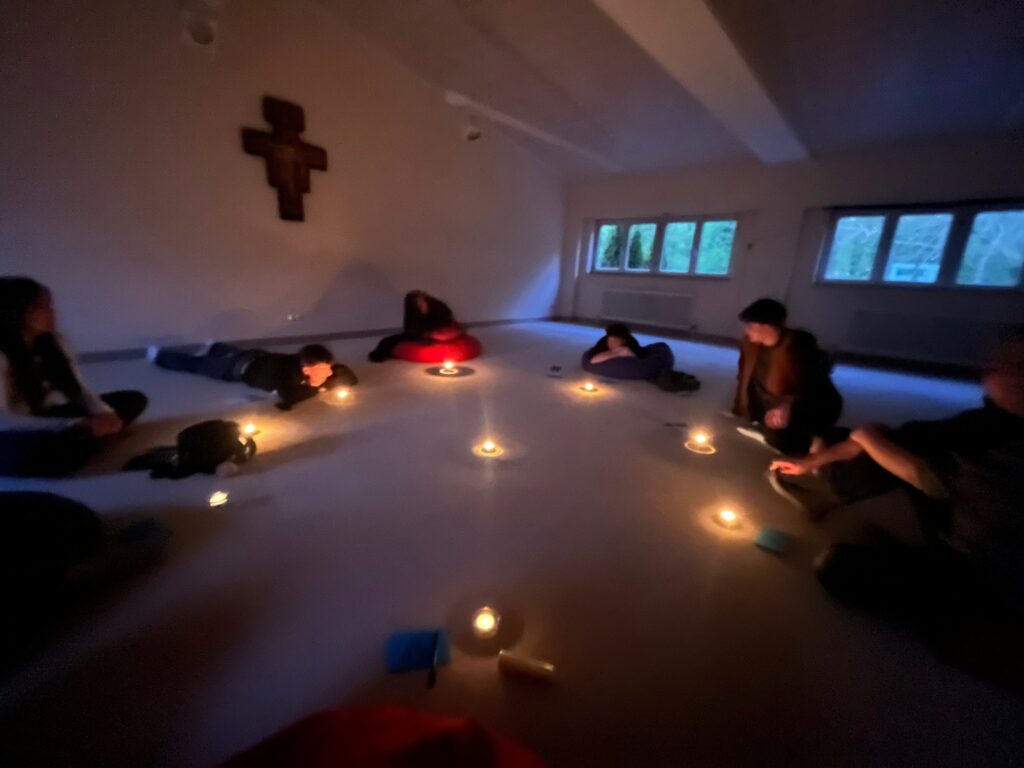 Menschen sitzen und liegen in einem schwach beleuchteten Raum um einen Kerzenkreis herum. An der Wand hängt ein Holzkreuz, und durch die Fenster ist das Grün draußen zu sehen.
