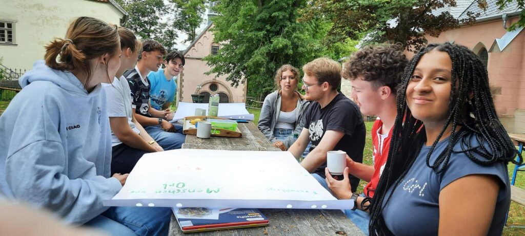 Eine Gruppe junger Leute sitzt um einen hölzernen Picknicktisch im Freien, unterhält sich und lächelt, auf dem Tisch stehen Notizbücher und Getränke. Im Hintergrund sind Bäume und Gebäude zu sehen.