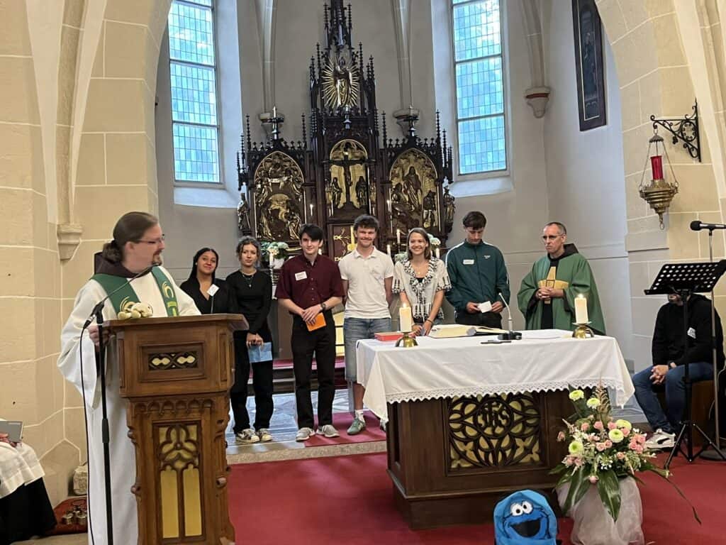 Ein Pfarrer spricht auf einem Podium in einer Kirche, während sieben Personen in der Nähe des Altars stehen; Kerzen und Blumen schmücken den Altar.
