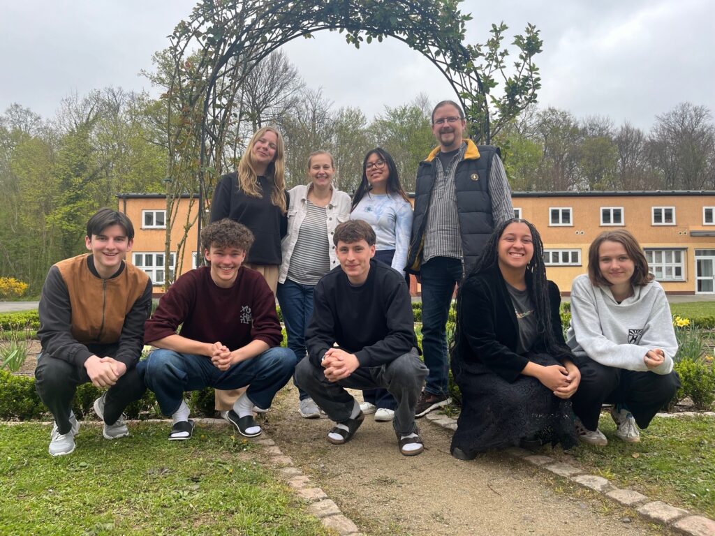 Eine Gruppe von neun Personen, darunter Erwachsene und Teenager, posiert an einem bewölkten Tag im Freien vor einem Gartenbogen und einem gelben Gebäude.