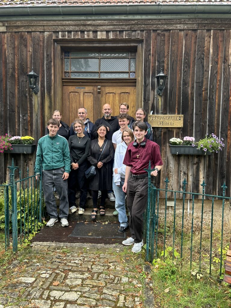 Eine Gruppe von zwölf Personen steht lächelnd vor einem rustikalen Holzgebäude mit einem Schild, umgeben von Blumen und einem Metallzaun.