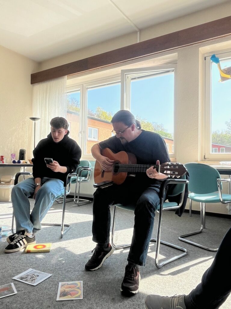 Zwei Personen sitzen auf Stühlen in einem sonnendurchfluteten Raum. Eine checkt ihr Telefon, während die andere Akustikgitarre spielt. Auf dem Boden liegen Kunstdrucke ausgebreitet.