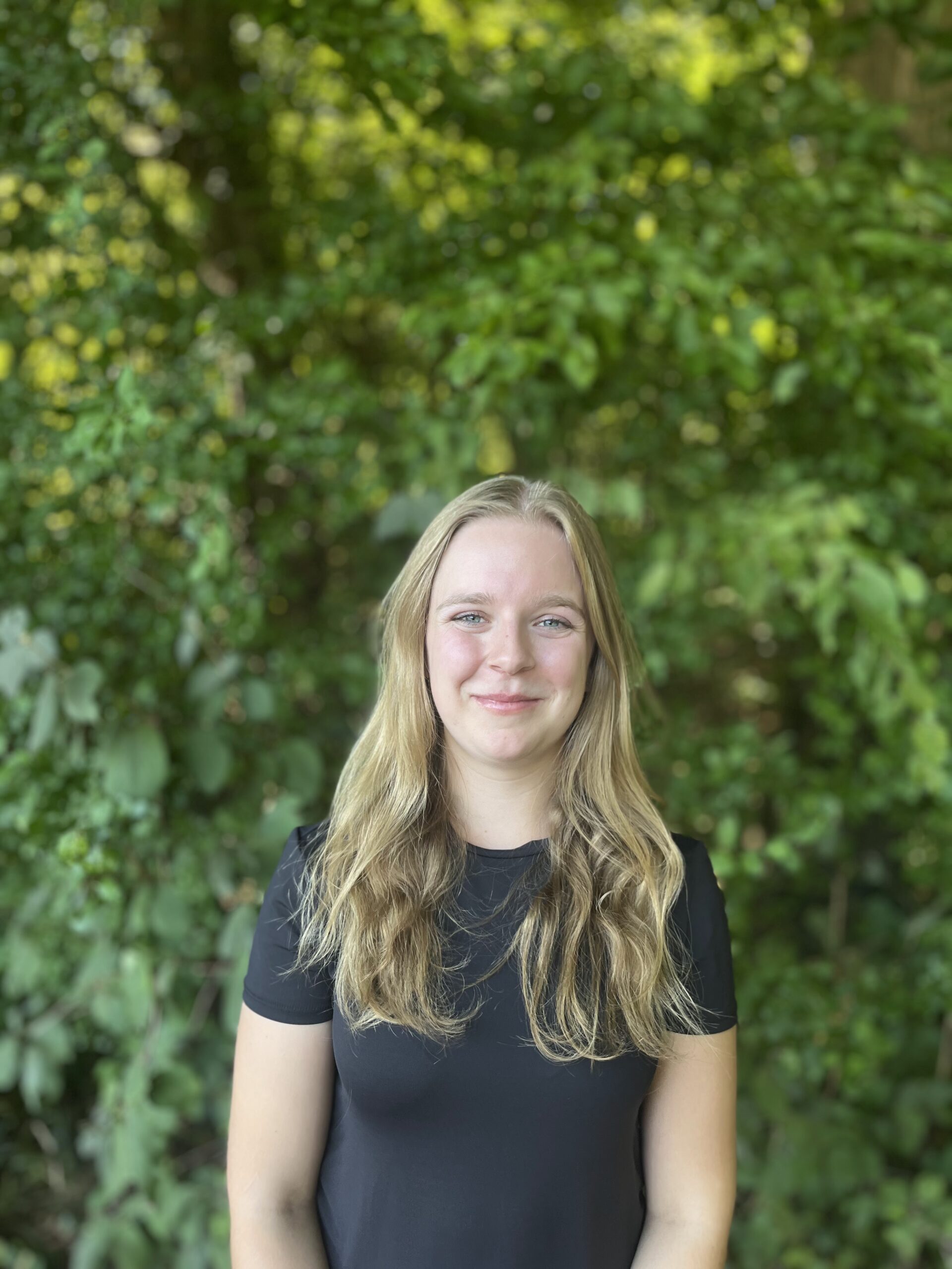 Eine Frau mit langen blonden Haaren und schwarzem Hemd steht im Freien vor grünem Laub, schaut in die Kamera und lächelt.
