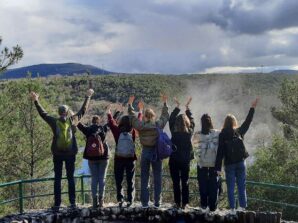 Eine Gruppe von Menschen mit Rucksäcken steht auf einem Felsvorsprung, blickt mit dem Rücken zur Kamera und streckt die Arme in Richtung einer malerischen Landschaft mit Hügeln, Bäumen und einem bewölkten Himmel aus.