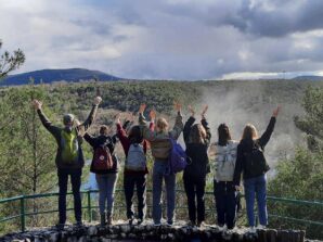 IMG_2287 Eine Gruppe von Menschen mit Rucksäcken steht auf einem Felsvorsprung, blickt mit dem Rücken zur Kamera und streckt die Arme in Richtung einer malerischen Landschaft mit Hügeln, Bäumen und einem bewölkten Himmel aus.